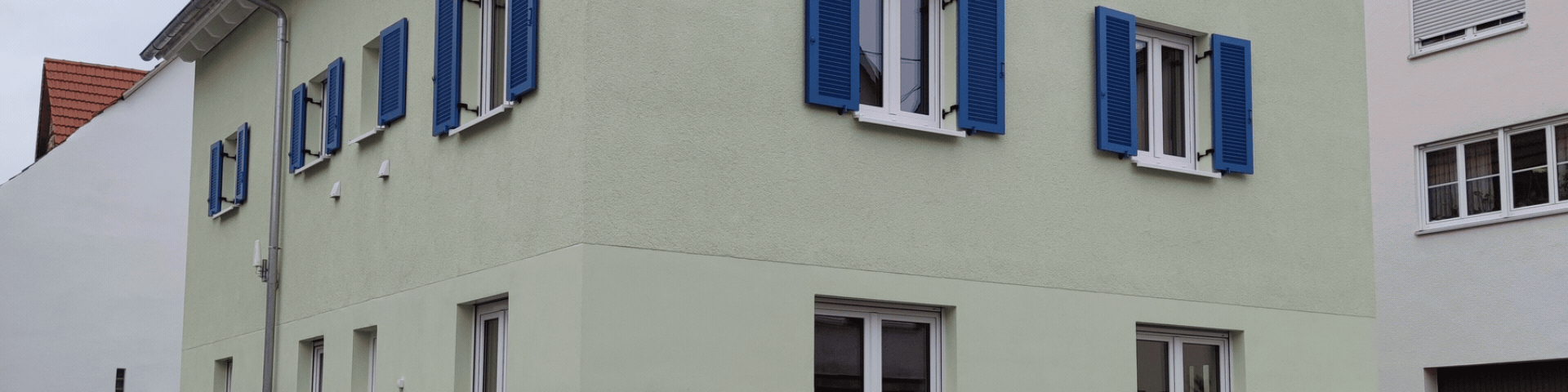 Bausweisen-1920x480 1 | Ehing Wohnbau GmbH | Ehing Wohnbau GmbH Two-story building with pale green walls and bright blue window shutters, white-framed windows along the facade.