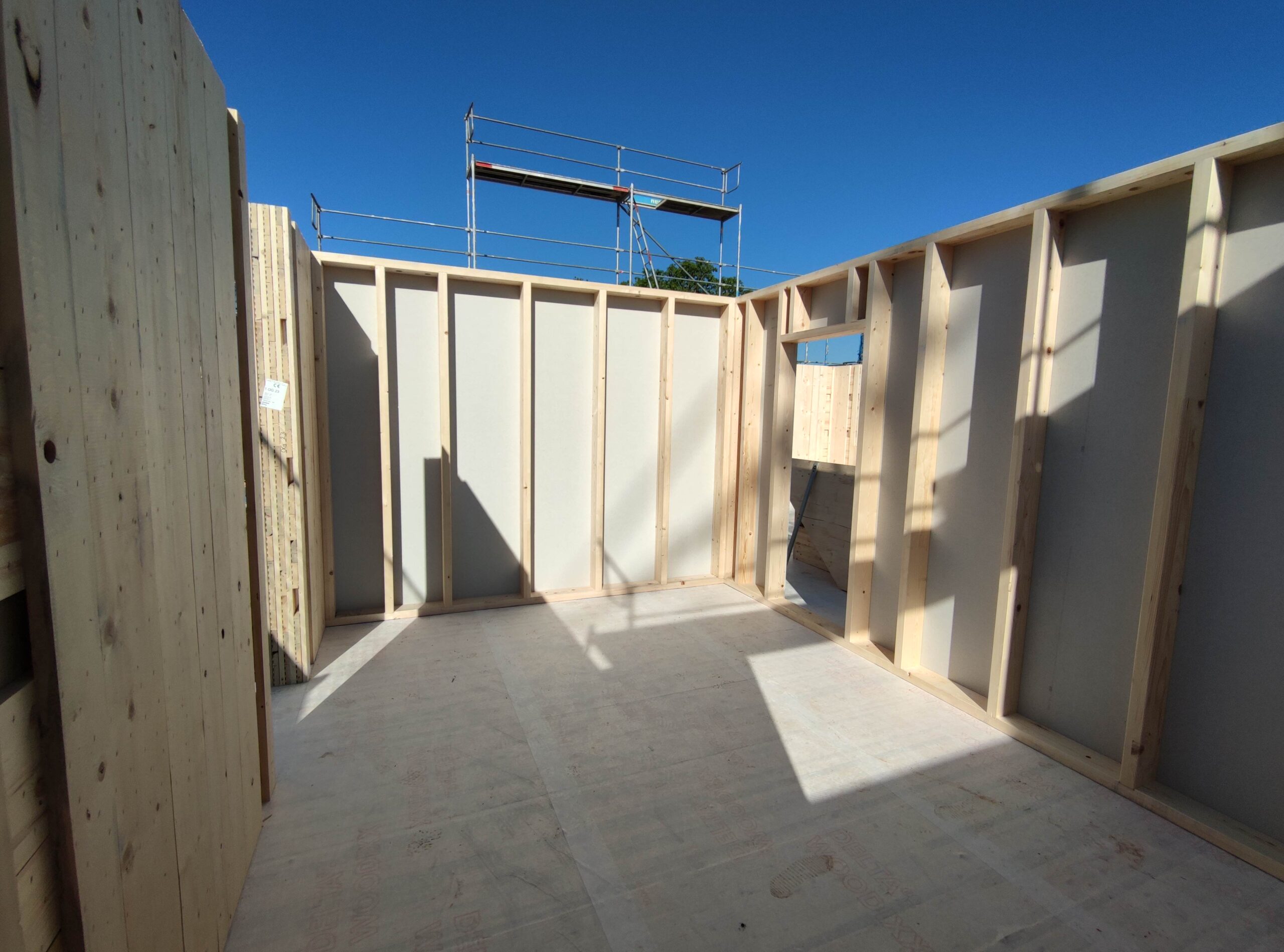 IMG_20240514_091212 | Ehing Wohnbau GmbH | Ehing Wohnbau GmbH Interior view of a room under construction with exposed wooden studs and framing, doorway opening, and blue sky visible outside.